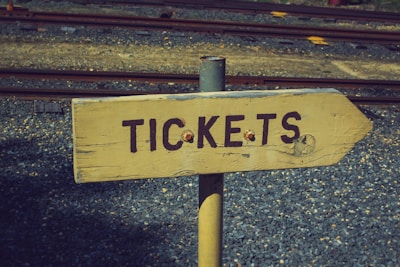 A wooden directional sign with the word 'TICKETS' painted in dark letters. The sign is mounted on a metal pole and set against a background of gravel and railway tracks.