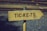A wooden directional sign with the word 'TICKETS' painted in dark letters. The sign is mounted on a metal pole and set against a background of gravel and railway tracks.