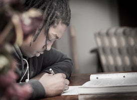A person with braided hair is focused on writing in a notebook while sitting at a table. Earphones are visible around their neck, and the environment appears to be calm and studious.