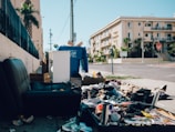 A street with litter and discarded items scattered along the sidewalk next to a dark leather chair. Boxes and a large blue recycling bin are visible, with an apartment building in the background. The area appears untidy and neglected.