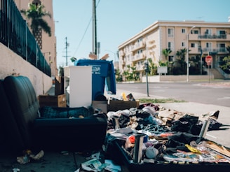 A street with litter and discarded items scattered along the sidewalk next to a dark leather chair. Boxes and a large blue recycling bin are visible, with an apartment building in the background. The area appears untidy and neglected.