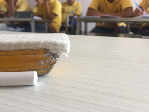 An old eraser and a piece of white chalk are lying on a classroom desk. In the background, there are students seated at desks, wearing yellow shirts, and engaged in some activities such as writing or reading.