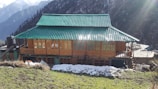 A rustic wooden house with a green tiled roof stands amidst a backdrop of snowy mountains and evergreen trees. Sunlight bathes the scene, casting rays across the structure, with patches of snow visible on the ground. The house features large windows with intricate wooden designs and a small staircase leading to the entrance.