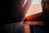 Strong angular architecture with dramatic lighting and deep shadows dominate the scene. The walls and surfaces are smooth, with a warm palette of browns and oranges. Geometric shapes create a play of light and shadow, casting sharp lines across the floor and walls.