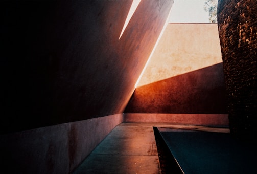 Strong angular architecture with dramatic lighting and deep shadows dominate the scene. The walls and surfaces are smooth, with a warm palette of browns and oranges. Geometric shapes create a play of light and shadow, casting sharp lines across the floor and walls.