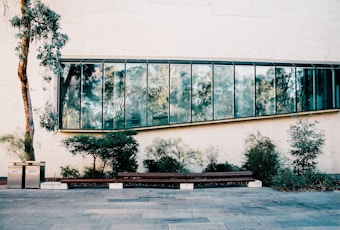 A modern architectural facade with large reflective glass windows set into a beige wall. In the foreground, there is a long wooden bench surrounded by small green shrubs and a single slender tree. Two waste bins labeled 'WASTE' and 'RECYCLE' are positioned near the tree. The scene is outdoors, with a paved area in front of the building.