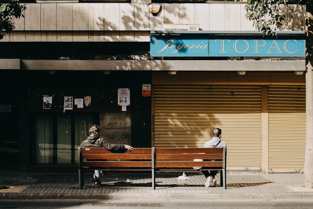 two persons sitting on bench near concrete buildings during daytime