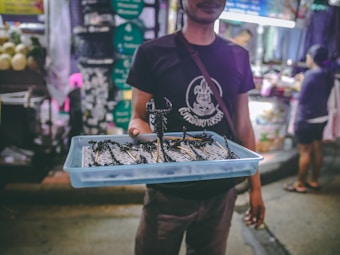 A person is holding a blue tray filled with skewered insects, likely on a busy street market. The background is blurry with vibrant lights and people shopping.