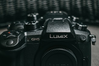 A close-up view of a black Lumix GH5 camera featuring various control dials and buttons, with a textured surface finish and the Lumix branding prominently displayed on top.