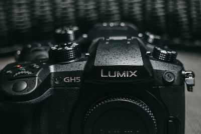 A close-up view of a black Lumix GH5 camera featuring various control dials and buttons, with a textured surface finish and the Lumix branding prominently displayed on top.