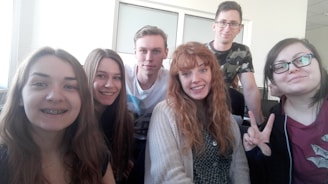 A group of six young individuals posing together for a cheerful photo. They are indoors, possibly in a classroom or office setting, sitting or standing close to each other with smiles. One person is showing a peace sign.