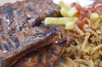 Close-up view of grilled ribs with char marks, served with golden fries topped with a drizzle of ketchup, and a side of crispy fried onions.