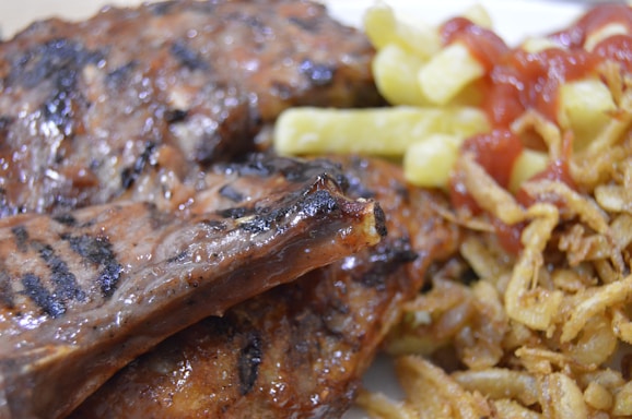 Close-up view of grilled ribs with char marks, served with golden fries topped with a drizzle of ketchup, and a side of crispy fried onions.