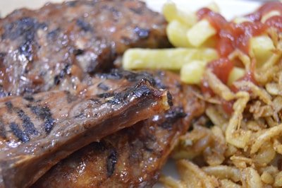 Close-up view of grilled ribs with char marks, served with golden fries topped with a drizzle of ketchup, and a side of crispy fried onions.