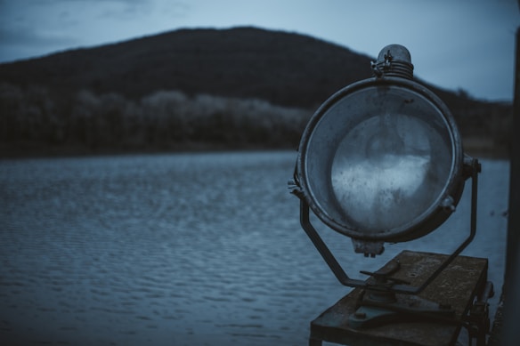 A large metal spotlight or floodlight is positioned in the foreground on a wooden platform near a body of water. The background features a blurred view of a hill or mountain under a dim, overcast sky. The overall atmosphere is calm and somewhat somber, with a cool color palette.