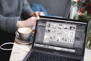 A person is using a laptop with a photo editing software open, featuring a grid of images on the screen. The scene is set in a cozy environment with a cup of coffee nearby on a white marble table. The blurred background suggests a casual or relaxed workspace.