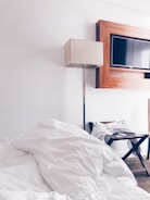 A neatly arranged hotel room features a large, unmade bed with white bedding in the foreground. A sleek floor lamp with a beige shade stands next to a modern, rectangular television mounted on the wall, framed with a wooden border. A luggage stand with a folded white robe is positioned beside the bed on the carpeted floor.