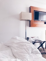 A neatly arranged hotel room features a large, unmade bed with white bedding in the foreground. A sleek floor lamp with a beige shade stands next to a modern, rectangular television mounted on the wall, framed with a wooden border. A luggage stand with a folded white robe is positioned beside the bed on the carpeted floor.