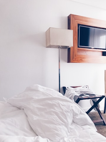 A neatly arranged hotel room features a large, unmade bed with white bedding in the foreground. A sleek floor lamp with a beige shade stands next to a modern, rectangular television mounted on the wall, framed with a wooden border. A luggage stand with a folded white robe is positioned beside the bed on the carpeted floor.