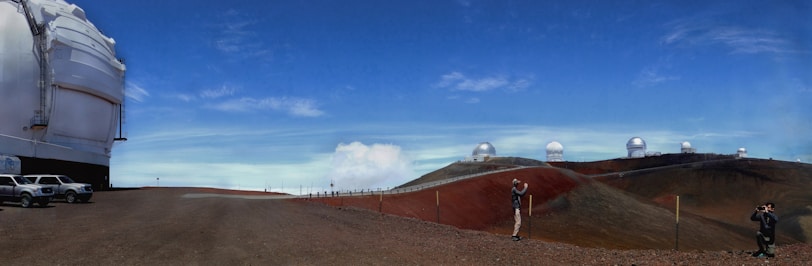 Multiple observatories with large domes are situated on a barren, rocky landscape under a clear blue sky. A couple of vehicles are parked nearby, and a few people are seen in the distance, possibly taking pictures.