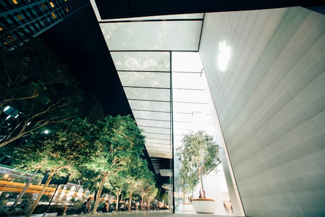 A modern architectural setting with tall glass walls reflecting the light. Several trees in planters line a pathway, creating a natural contrast against the urban surroundings. The scene suggests a high-tech and environmentally conscious design.