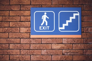 A blue sign with a white figure walking towards the word 'EXIT' accompanied by a graphic of stairs is mounted on a brick wall.