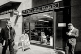A barbershop with a sign reading 'Pall Mall Barbers' is pictured. Two men in business attire are outside; one is smiling and walking away while the other is leaning against the wall, looking at a mobile phone. Inside, another person is seated in a barber chair.