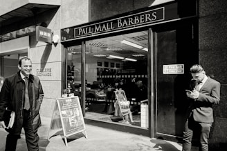 A barbershop with a sign reading 'Pall Mall Barbers' is pictured. Two men in business attire are outside; one is smiling and walking away while the other is leaning against the wall, looking at a mobile phone. Inside, another person is seated in a barber chair.