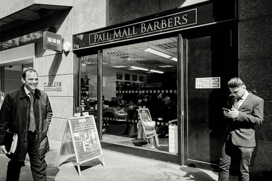 A barbershop with a sign reading 'Pall Mall Barbers' is pictured. Two men in business attire are outside; one is smiling and walking away while the other is leaning against the wall, looking at a mobile phone. Inside, another person is seated in a barber chair.