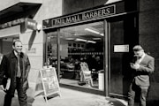 A barbershop with a sign reading 'Pall Mall Barbers' is pictured. Two men in business attire are outside; one is smiling and walking away while the other is leaning against the wall, looking at a mobile phone. Inside, another person is seated in a barber chair.
