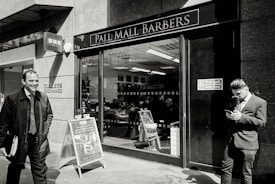 A barbershop with a sign reading 'Pall Mall Barbers' is pictured. Two men in business attire are outside; one is smiling and walking away while the other is leaning against the wall, looking at a mobile phone. Inside, another person is seated in a barber chair.