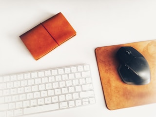 A neatly arranged workspace with a white keyboard, a leather-bound notebook secured with a red elastic band, and a black computer mouse on a brown leather mouse pad. The items are placed against a clean, light-colored background.