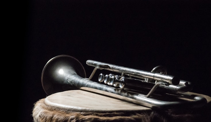 A silver trumpet rests on a circular wooden drum covered with animal skin, set against a dark, moody background. The instrument's metallic sheen is subtly highlighted by light, creating a contrast with the shadowy surroundings.
