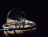A shiny brass musical instrument, specifically a French horn, rests against a dark background. The intricate details and curves of the metal are emphasized by the lighting, which creates reflections and highlights the craftsmanship. The surface on which the instrument is placed has a textured, natural appearance.