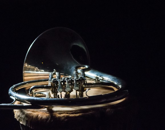 A shiny brass musical instrument, specifically a French horn, rests against a dark background. The intricate details and curves of the metal are emphasized by the lighting, which creates reflections and highlights the craftsmanship. The surface on which the instrument is placed has a textured, natural appearance.