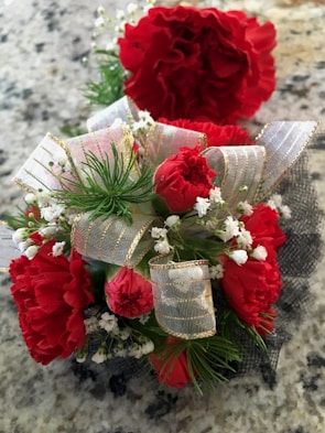 A close-up view of a corsage featuring bright red carnations adorned with small white flowers and decorative ribbons on a speckled granite surface.