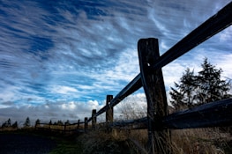 A wooden fence stretches diagonally across a rural landscape under a dramatic sky filled with streaked clouds. Tall grasses and silhouetted trees are visible in the background, adding to the natural, serene scene.