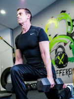 A man wearing a black athletic shirt and pants is performing a lunge exercise while holding a dumbbell in each hand. The setting appears to be a gym, indicated by the presence of gym equipment like tires and resistance bands. Vibrant graffiti-style art with green and yellow accents decorates the wall behind him.
