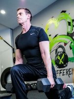 A man wearing a black athletic shirt and pants is performing a lunge exercise while holding a dumbbell in each hand. The setting appears to be a gym, indicated by the presence of gym equipment like tires and resistance bands. Vibrant graffiti-style art with green and yellow accents decorates the wall behind him.