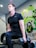A man wearing a black athletic shirt and pants is performing a lunge exercise while holding a dumbbell in each hand. The setting appears to be a gym, indicated by the presence of gym equipment like tires and resistance bands. Vibrant graffiti-style art with green and yellow accents decorates the wall behind him.
