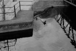 A black and white image of a concrete platform with the number 1928 etched on its side, adjacent to a body of water. Reflection of railings and a tree are visible in the water, giving it a somewhat tranquil yet eerie feel. The water surface shows gentle ripples with a submerged log or branch partially visible.