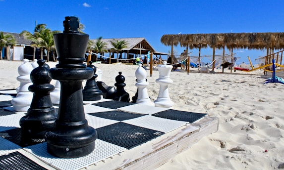 A large outdoor chess set is placed on a sandy beach with rows of palm trees and thatched-roof structures in the background. The chess pieces are oversized and made of plastic, consisting of black and white pieces on a checkerboard pattern. Hammocks and lounge chairs are visible near the huts, suggesting a relaxing beach environment.