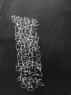 A vertical sequence of white graffiti-like text is scribbled on a dark surface, possibly a wall or a chalkboard. The text includes several words, some recognizable and some not, with an artistic and somewhat chaotic arrangement and style.