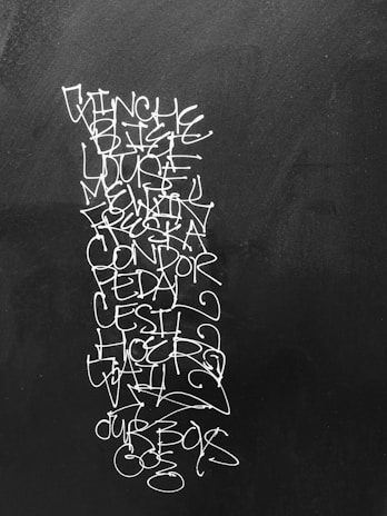 A vertical sequence of white graffiti-like text is scribbled on a dark surface, possibly a wall or a chalkboard. The text includes several words, some recognizable and some not, with an artistic and somewhat chaotic arrangement and style.