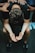 A person is sitting with their head bent forward, arms resting on their knees, and palms open in a pleading or contemplative gesture. Multiple hands are laid on their back, suggesting support or comfort from others around them. The atmosphere feels solemn and empathetic.