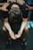 A person is sitting with their head bent forward, arms resting on their knees, and palms open in a pleading or contemplative gesture. Multiple hands are laid on their back, suggesting support or comfort from others around them. The atmosphere feels solemn and empathetic.