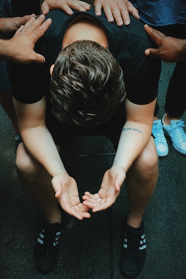 A person is sitting with their head bent forward, arms resting on their knees, and palms open in a pleading or contemplative gesture. Multiple hands are laid on their back, suggesting support or comfort from others around them. The atmosphere feels solemn and empathetic.