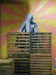 A mural of a historical figure is painted on a brick wall with a colorful background of radiating pink and yellow rays. Various small icons, like pineapples and bicycles, are scattered across the wall. Wooden pallets are stacked in front of the mural, partially obscuring the figure.