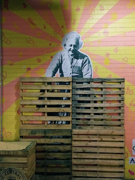 A mural of a historical figure is painted on a brick wall with a colorful background of radiating pink and yellow rays. Various small icons, like pineapples and bicycles, are scattered across the wall. Wooden pallets are stacked in front of the mural, partially obscuring the figure.