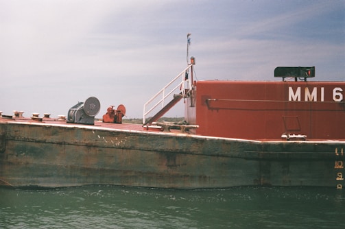 A weathered barge is floating on calm water. The vessel has a rusty appearance, with visible metal wear and discoloration. Its surface is equipped with some machinery, including a winch or pulley system, and a red-hued cabin or structure. A simple railing leads up to the cabin area. The sky overhead is slightly overcast, adding a muted tone to the scene.